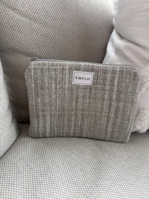 Beige tygnecessär från Emelo - Snygg beige necessär från Emelo i tyg med diskret randigt mönster. Väskan har dragkedja upptill och en ljus insida. Perfekt för småprylar, smink eller som organiserare i större väska. Enkel och stilren design som passar till det mesta. Mått ca 20x15 cm