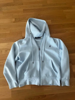 Ljusblå hoodie från Polo Ralph Lauren - Snygg ljusblå hoodie från Polo Ralph Lauren med dragkedja framtill, två fickor och justerbar huva med snören. Klassisk logga broderad på bröstet. Tillverkad i mjukt bomullsmaterial som känns skönt mot huden.