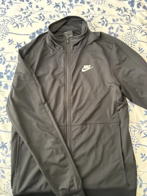 Svart träningsjacka från Nike - Snygg svart träningsjacka från Nike med dragkedja framtill och hög krage. Jackan har två sidofickor och en vit Nike-logga på bröstet. Perfekt för träning eller chill. Tillverkad i ett mjukt och stretchigt material.