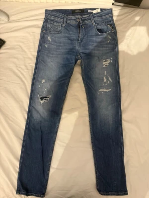 Replay jeans - Säljer ett par blå jeans från Replay, modell Anbass. Jeansen har slitna detaljer och en klassisk femficksdesign. De är tillverkade i jeansmaterial och har en normal passform med dragkedja och knapp i midjan. Perfekta för en avslappnad och trendig look. Dessa kostar 1600 kronor nya.