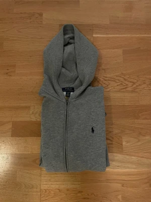 Grå hoodie från Polo Ralph Lauren - Snygg grå hoodie från Polo Ralph Lauren med dragkedja framtill och klassisk huva. Diskret logga broderad på bröstet. Tillverkad i mjukt material som känns skönt mot huden. Perfekt för en avslappnad och stilren look.