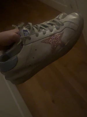 Golden Goose vita sneakers med glitterstjärna - Säljer ett par vita Golden Goose sneakers med en rosa glitterstjärna på sidan och ljusblå detaljer vid hälen. Skorna har klassisk låg profil, gråa skosnören och är tillverkade i läder. Perfekt för dig som vill ha något unikt och trendigt. Ganska utslitna därav priset! OBS lådan och påsen kommer med