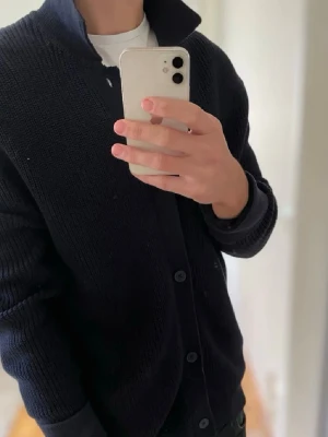 Mörkblå stickad kofta med krage - Säljer en stilren mörkblå stickad kofta med hög krage och knäppning framtill. Koftan har ribbade muddar vid ärmsluten och en klassisk, rak passform. Perfekt att slänga över en t-shirt för en avslappnad men snygg look.