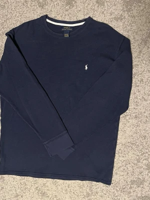 Mörkblå långärmad tröja Polo Ralph Lauren - Mörkblå långärmad tröja från Polo Ralph Lauren med klassisk vit broderad logga på bröstet. Tröjan har rund halsringning och är tillverkad i ett mjukt, strukturerat material som känns skönt mot huden. Perfekt för en stilren och avslappnad look.