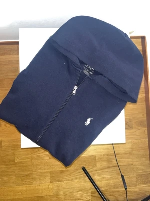 Mörkblå hoodie från Ralph Lauren - Snygg mörkblå hoodie från Ralph Lauren med dragkedja framtill och klassisk vit logga på bröstet. Hoodien har huva och är tillverkad i mjukt bomullsmaterial som känns skönt mot huden. Perfekt för en avslappnad och stilren look.
