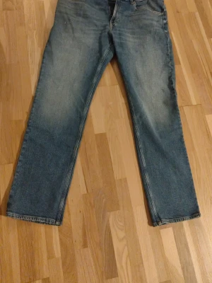 Blå raka jeans från Tommy Hilfiger - Säljer ett par klassiska blå jeans från Tommy Hilfiger med rak passform och femficksmodell. Jeansen har en snygg tvätt med lätt slitning framtill och bakfickor med diskret logga. Helt nya har aldrig blivit använda.