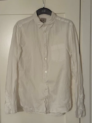 Vit linneskjorta från H&M - Klassisk vit skjorta från H&M i regular fit med lång ärm och bröstficka. Tillverkad i svalt linne, perfekt för en clean och stilren look. Skjortan har knappar framtill och enkel krage. Nästan helt ny