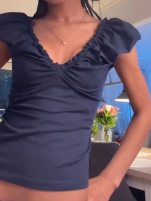 Mörkblå topp med volang och v-ringning - Snygg mörkblå topp med v-ringning och volangdetaljer längs halsen. Toppen har korta puffärmar och sitter figurnära. Var helt slutsåld och nu inte säljs längre. Andvänd 1 gång 