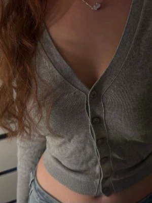 Grå cropped kofta med knappar - Snygg grå cropped kofta med v-ringning och långa ärmar. Koftan har ribbade muddar och knäppning framtill med fem knappar. Perfekt att styla med jeans eller kjol för en trendig look.