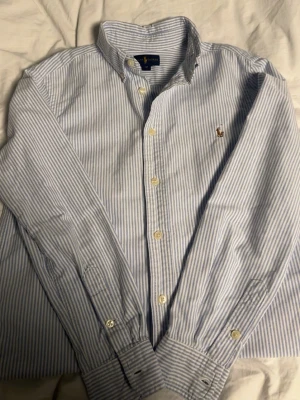 Blårandig skjorta från Polo Ralph Lauren - Klassisk blårandig skjorta från Polo Ralph Lauren med button-down krage och lång ärm. Skjortan har vita och ljusblå smala ränder, broderad logga på bröstet och pärlemorsknappar. Tillverkad i bomull för en skön och luftig känsla.