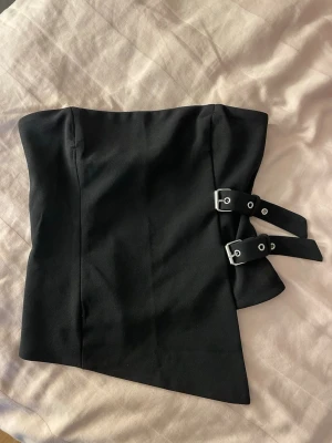 Svart asymmetrisk korsett med spännen - Säljer en svart korsett med asymmetrisk nederkant och två dekorativa spännen i silver på sidan. Korsetten är axelbandslös och har en stilren, modern look. Materialet känns kraftigt och håller formen snyggt. Perfekt för dig som vill ha en edgy och trendig stil.