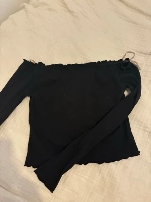 Svart offshoulder topp med långa ärmar - Trendig svart offshoulder topp med långa ärmar och ribbad struktur. Toppen har vågig kant upptill och nedtill för en extra snygg detalj. Perfekt att styla med jeans eller kjol för en cool look. Forfarande i bra skick och använts fåtal gånger. Kom med egna bud eller vid intresse