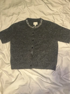 Grå kortärmad kofta med pärlor H&M - Snygg grå stickad kofta från H&M med korta ärmar och knappar framtill. Rund halsringning dekorerad med glittrande pärlor och stenar. Mjuk stickad kvalitet. Den sticker ej. 💗Är i storlek M, men känns som storlek S