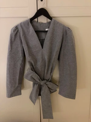 Grå omlottblus med knytband - Stilren grå omlottblus med djup V-ringning och breda knytband i midjan. Blusen har puffiga axlar och långa ärmar för en trendig siluett men kan även stylas på massa olika sätt, med eller utan band 🎀 