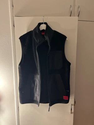 Svart fleeceväst från Hugo - Snygg svart fleeceväst från Hugo med dragkedja framtill. Västen har en mjuk, lurvig yta och detaljer i slätare tyg på framsidan. Perfekt för lager på lager och enkel att matcha med olika outfits.