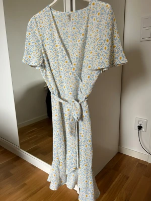 Blommig omlottklänning med volang - Söt långklänning i ljusblått och vitt med gula blommor. Omlottmodell med knytband i midjan, korta ärmar och volangkant nedtill. Perfekt för sommarens alla tillfällen.