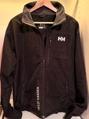 Svart vindjacka från Helly Hansen - Svart vindjacka från Helly Hansen med vit HH-logga på bröstet och HELLY HANSEN-tryck vid dragkedjan. Jackan har hög krage med grå insida. Storlek M
