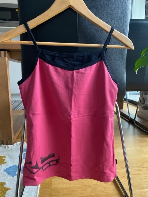 Rosa linnetopp med svarta detaljer - Snygg rosa linnetopp med tunna svarta axelband och svart kant vid halsen. Toppen har ett tryck i svart nere till vänster och är figurnära i modellen. Perfekt för dig som gillar sportig och enkel stil.