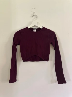 Vinröd ribbad croppad topp - Trendig vinröd croppad topp med lång ärm och rund hals. Toppen är ribbad för en snygg struktur och har en lätt böjd nederkant framtill. Perfekt att styla med höga jeans eller kjol.