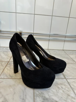 Svarta platåpumps med hög klack 39 - Snygga svarta pumps med rund tå och rejäl platåsula. Skorna har hög klack och är klädda i ett sammetsliknande material som ger en lyxig känsla. Perfekta för dig som vill ha extra höjd och en klassisk look.