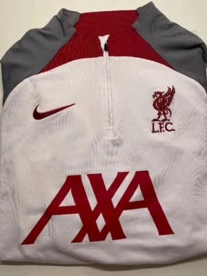 Liverpool Nike träningströja  - Säljer en snygg Liverpool FC träningströja från Nike i vitt med röda och grå detaljer. Tröjan har halv dragkedja, klubbmärke och Nike-logga på bröstet samt AXA-tryck framtill. Tillverkad i funktionsmaterial som andas, perfekt för träning.