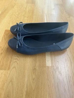 Svarta ballerinaskor från Geox - Snygga svarta ballerinaskor från Geox med rund tå och dekorativ rosett framtill. Skorna är i ett slätt material och har diskreta sömmar längs kanterna. Perfekta för en stilren och enkel look.