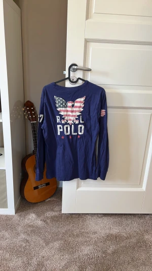 Blå långärmad topp från Polo - Snygg marinblå långärmad topp från Polo med stort tryck av en örn och amerikanska flaggan på ryggen. Trycket har texten 'RL POLO' och detaljer på ärmarna. Perfekt för dig som gillar sportig och avslappnad stil.