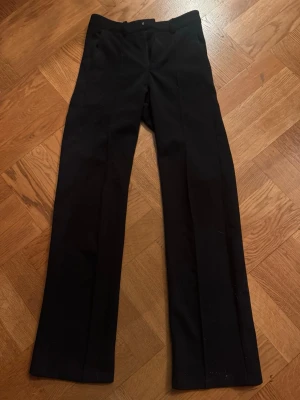 Svarta raka kostymbyxor - Snygga svarta kostymbyxor med rak passform och pressveck. Klassisk midja och diskreta fickor framtill. Perfekta för en clean och stilren look. Materialet ser ut att vara en mjuk och lite stretchig blandning, vilket gör dem bekväma hela dagen.