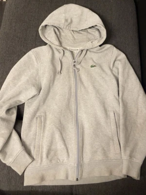 Grå hoodie från Lacoste med dragkedja - Snygg ljusgrå hoodie från Lacoste i classic fit med dragkedja framtill och två fickor. Ikoniska gröna krokodilloggan på bröstet. Tillverkad i mjukt bomullsmaterial och har justerbar huva med snören. Perfekt för en chill och stilren look.