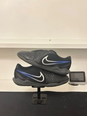 Nike svart/blå fotbollsskor - Säljer ett par svarta fotbollsskor från Nike med blå och vita detaljer. Skorna har en låg profil och är tillverkade i syntetmaterial med en vågig struktur på ovansidan. Den klassiska Nike-loggan syns tydligt på sidan och hälen har en unik symbol.