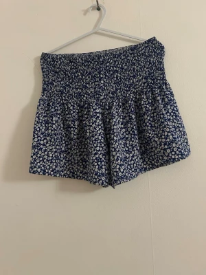 Blommiga blå shorts med hög midja - Supersöta blå shorts med vitt blommönster och smockad, hög midja. Luftig och flowy modell som ger en avslappnad vibe. Perfekta för varma dagar och lätta att matcha med en enkel topp.