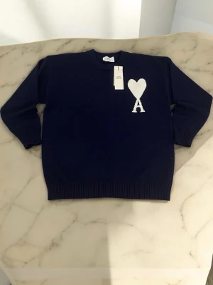 Mörkblå stickad tröja Ami Paris - Mörkblå stickad tröja från Ami Paris med vit hjärtlogga och bokstaven A på bröstet. Tröjan har rund halsringning, långa ärmar och ribbade muddar vid ärmslut och nederkant. Perfekt för dig som gillar stilrena och trendiga plagg.