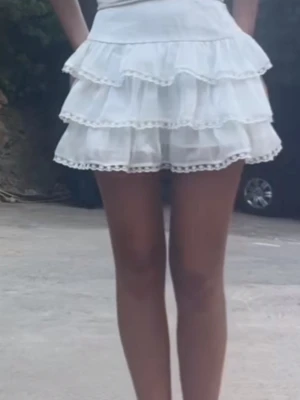 Vit volangkjol med spetsdetaljer - Supersöt vit kortkjol med flera lager volanger och fina spetsdetaljer längs kanterna. Kjolen har en hög midja och ger en luftig och romantisk känsla, perfekt för varma dagar.