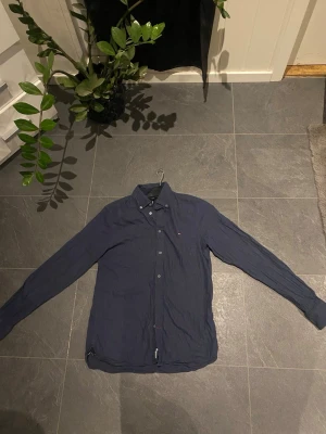 Mörkblå skjorta från Tommy Hilfiger - Klassisk mörkblå skjorta från Tommy Hilfiger i mjuk bomull. Skjortan har långa ärmar, knappar framtill och en stilren krage. Perfekt för dig som gillar en enkel men snygg look.
