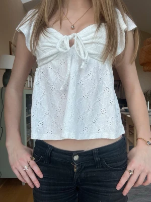 Vit broderad blus - Söt vit blus i bomull med broderade detaljer och korta ärmar. Toppen har en v-ringning med knyt framtill och lätt volang nedtill. Perfekt till jeans eller kjol för en fräsch look.