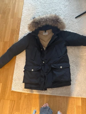 Svart dunjacka med päls från Woolrich - Säljer en svart dunjacka från Woolrich med beige quiltad insida och stor huva med lyxig pälsdetalj. Jackan har två stora fickor med lock och knappar framtill, perfekt för kalla dagar. Klassisk och tidlös design som håller dig varm.