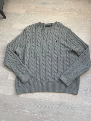 Grå kabelstickad tröja Polo Ralph Lauren - Snygg grå kabelstickad tröja från Polo Ralph Lauren med klassisk rund halsringning och broderad marinblå logga på bröstet. Tröjan har långa ärmar och ribbade muddar vid ärmslut och nederkant. Perfekt för lager på lager och stilren vardagslook.