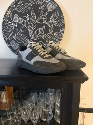 Grå sneakers från Axel Arigato - Snygga sneakers från Axel Arigato i olika grå nyanser med svarta detaljer och vita snören. Skorna har en chunky sula och är tillverkade i mocka och textil. Märket syns i guld på sidan. Perfekta för dig som gillar stilrena och moderna sneakers.