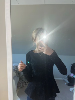 Svart peplum topp med lång ärm - Säljer en svart peplum topp med lång ärm och rund halsringning. Toppen har en figurnära passform upptill och en volang nedtill som ger en snygg siluett. Perfekt att styla med jeans eller kjol för en trendig look.