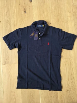 Marinblå pikétröja från Ralph Lauren - Klassisk marinblå pikétröja från Ralph Lauren med korta ärmar och krage. Tröjan har två knappar vid halsen och den ikoniska röda logotypen broderad på bröstet. Tillverkad i mjuk bomull som känns skön mot huden.