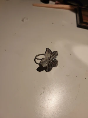Silverfärgad blomring i metall - Unik ring i silverfärgad metall med en stor blomma som dekoration. Blomman har detaljerade blad och små pärlliknande detaljer i mitten. Perfekt för dig som vill ha ett statement-smycke med naturinspirerad design.