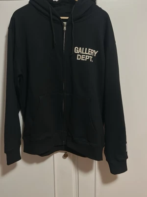 Svart hoodie från Gallery Dept. - Säljer en svart hoodie från Gallery Dept. med dragkedja framtill och vit logga på bröstet. Hoodien har huva med dragsko och en stor magficka. Tillverkad i mjukt material som känns skönt mot huden. Perfekt för en avslappnad streetwear-look.
