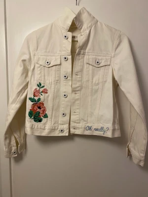 Vit jeansjacka med broderi från H&M - Snygg vit jeansjacka från H&M med broderade blommor i rosa och grönt på ena sidan samt texten 'Oh, really?' broderad vid nederkanten. Klassisk modell med bröstfickor och silverfärgade knappar. Perfekt för dig som vill sticka ut lite extra.