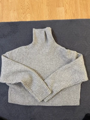 Grå ribbstickad polotröja - Säljer en grå ribbstickad polotröja med hög krage och långa ärmar. Tröjan har en boxig, lite croppad passform och är perfekt för lager på lager. Materialet känns mjukt och stretchigt, vilket gör den superbekväm.