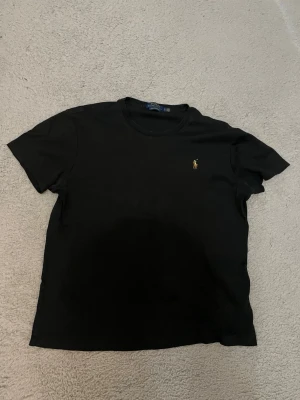 Svart t-shirt från Polo Ralph Lauren - Säljer en svart t-shirt från Polo Ralph Lauren i custom slim fit. Klassisk design med korta ärmar och den ikoniska lilla broderade loggan på bröstet. Tillverkad i mjuk bomull som känns skön mot huden. Perfekt till jeans eller shorts.