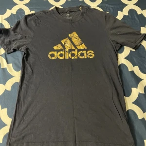 Mörkblå Adidas t-shirt med gul logga - Snygg mörkblå t-shirt från Adidas med stor gul logga och grafiskt mönster på bröstet. Klassisk rund halsringning och korta ärmar. Perfekt för en sportig och avslappnad stil.