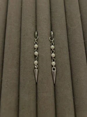 Silvriga örhängen med pärlor och spikes - Säljer ett par trendiga örhängen i silverfärg med små vita pärlor på rad och spetsiga spikes längst ner. Örhängena har en cool kedjedesign och passar perfekt för dig som gillar en edgy stil. Materialet ser ut att vara metall och pärlor.