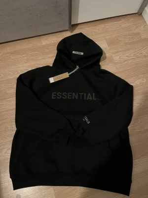 Svart Essentials Fear of God hoodie - Säljer en svart hoodie från Fear of God Essentials med stor huva och ficka framtill. Hoodien har tryckt ESSENTIALS-text på bröstet och diskret logga på ärmen. Tillverkad i mjukt bomullsmaterial för en clean och stilren look.