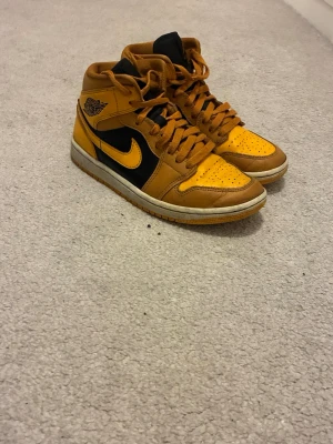Nike Air Jordan 1 Mid gul/svart - Säljer ett par Nike Air Jordan 1 Mid sneakers i gul och svart med klassisk vinge-logga på sidan och svart swoosh. Skorna har en vit mellansula och gult yttersula, samt perforerad tåbox och svarta detaljer på plösen. Tillverkade i läder och textil för en ikonisk look.