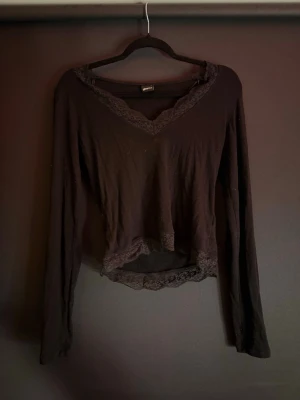 Svart långärmad topp från Gina Tricot - Svart långärmad topp från Gina Tricot med v-ringning och spetsdetaljer vid halsringning och nederkant. Toppen har en något croppad passform och är tillverkad i mjukt material som känns skönt mot huden. Perfekt för att lyfta vilken outfit som helst.